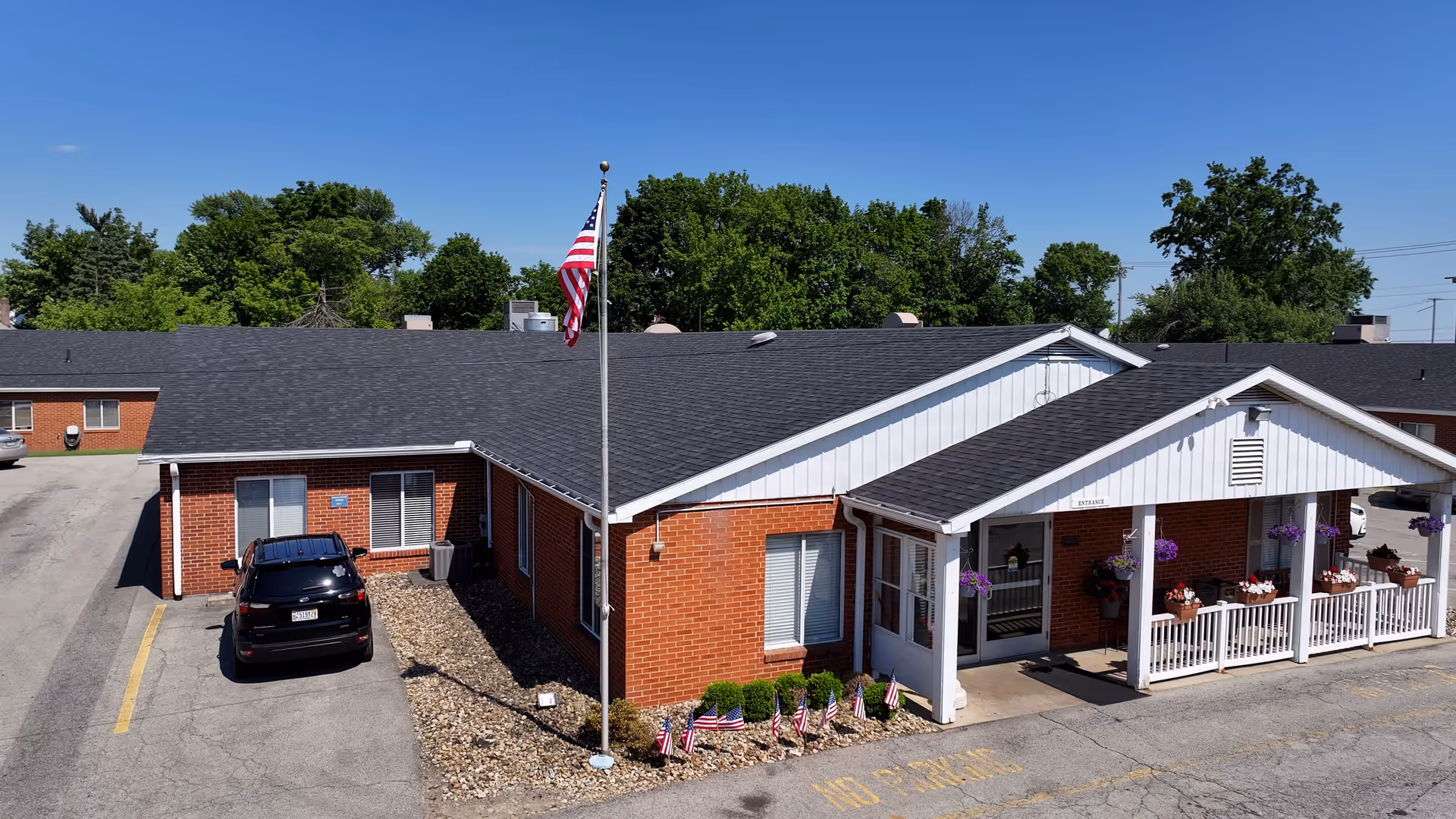Exterior view of a single-story brick building with a black roof, an American flag on a flagpole in front, and a covered entrance decorated with hanging flower baskets and potted plants. A black car is parked near the building, and there are small American flags planted in the landscaping along the front.