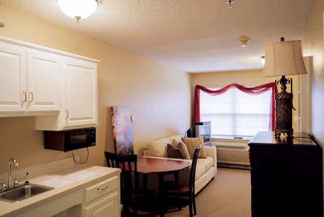 Interior view of a small living space at Winkler Court featuring a kitchenette with white cabinets and a sink on the left, a round wooden table with two chairs, a beige sofa with cushions, a TV on a stand near a window with red curtains, and a dark wooden dresser with a lamp on top on the right.