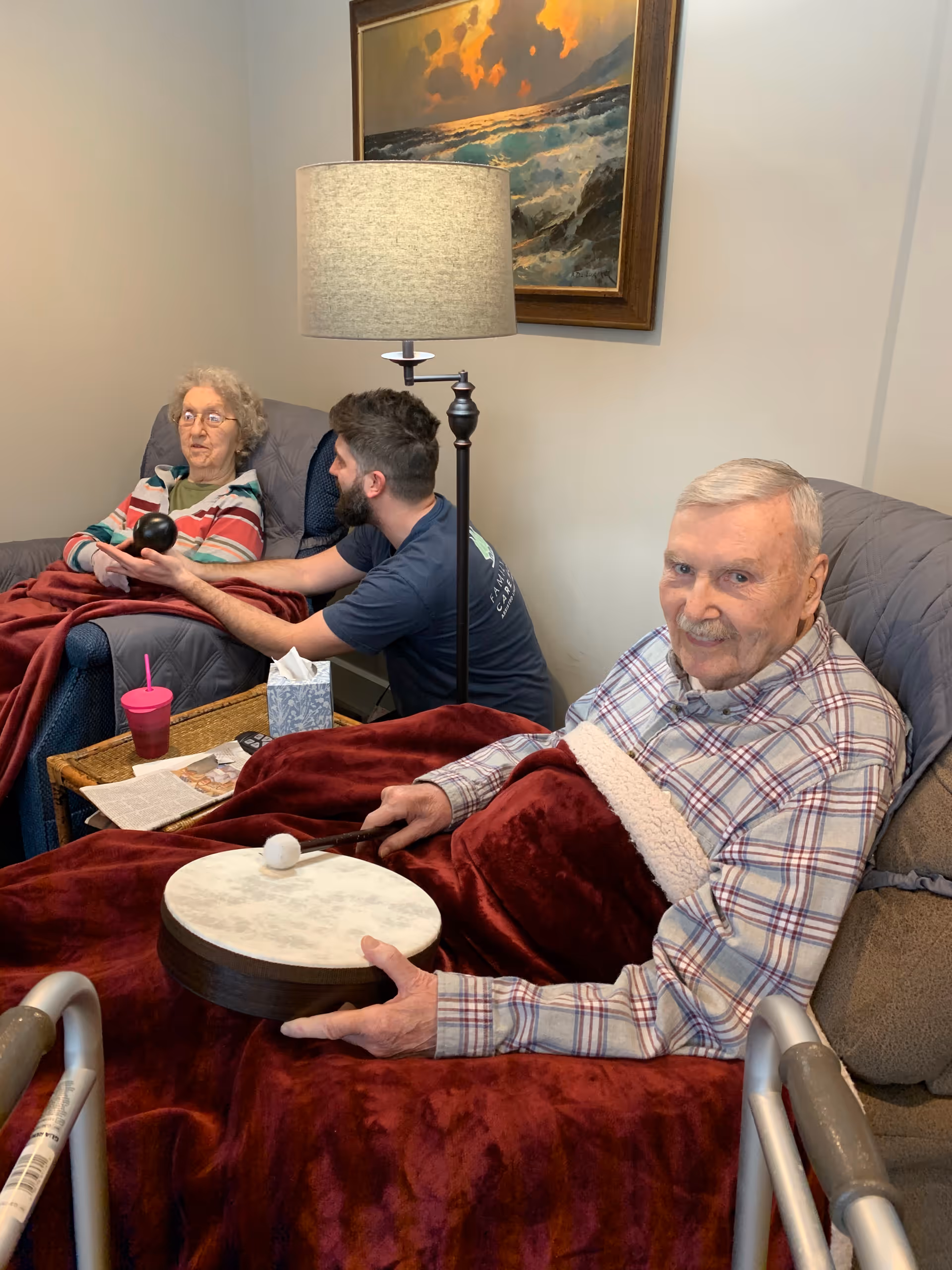 Two elderly residents seated in recliners in a cozy living room with a caregiver between them; one resident holds a small drum and mallet.