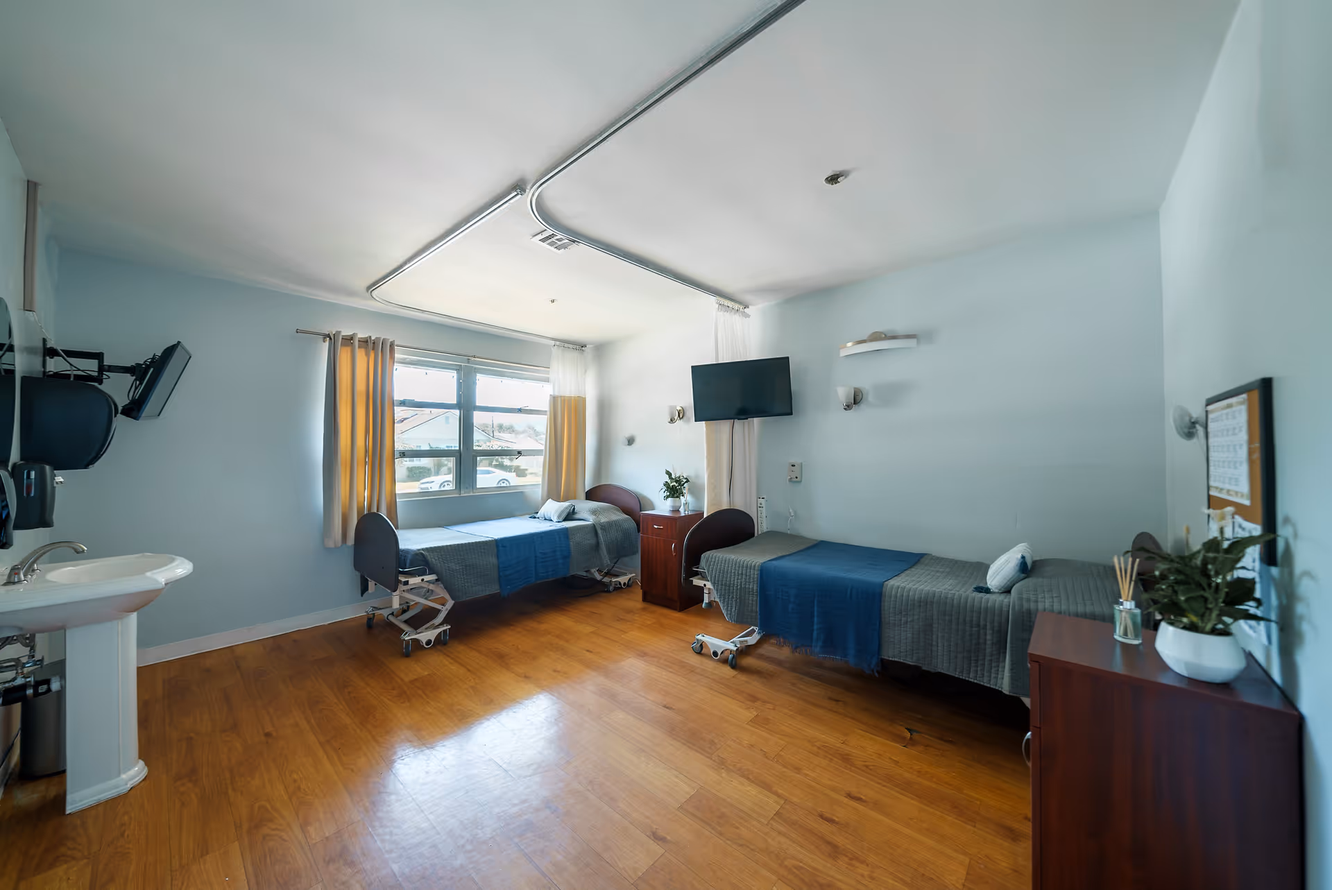 Shared patient bedroom in a rehabilitation facility with two hospital beds, a wall-mounted TV, a sink, and a window with curtains.