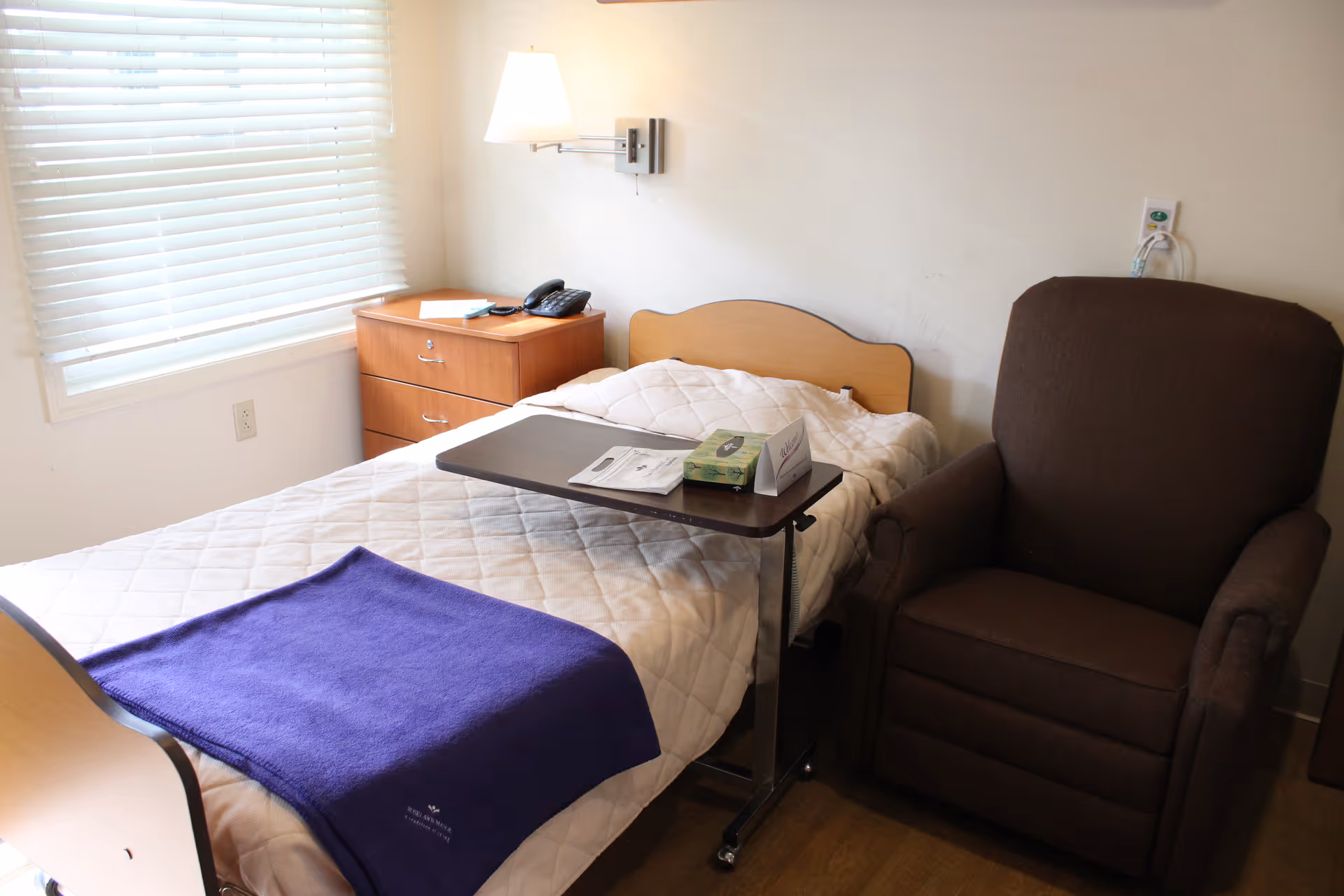 A small bedroom with a single bed covered in a white quilt and a purple blanket at the foot. Next to the bed is a brown recliner chair and a wooden nightstand with a telephone and some papers. A window with white blinds is on the left wall, and a wall-mounted lamp is above the nightstand. A small overbed table with tissues and papers is positioned over the bed.