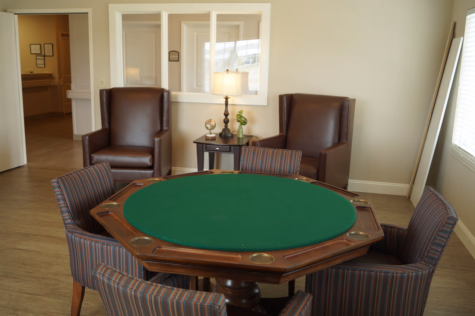 A cozy interior room with a green felt octagonal game table surrounded by four striped upholstered chairs. Behind the table are two brown leather armchairs with a small side table between them holding a lamp, a small globe, and a plant. The room has light-colored walls and wood flooring, with a window on the right side letting in natural light.