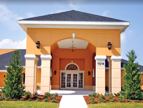 Front exterior view of a building with a large covered entrance supported by four columns, orange and beige walls, a chandelier hanging from the ceiling of the entrance, and the number 11214 displayed on one of the columns. There are small landscaped bushes and trees on either side of the walkway leading to the entrance.