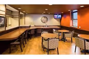 Interior view of a common area with wooden flooring and ceiling, featuring a long counter with chairs along one wall and several round tables with cushioned chairs. A television is mounted on the wall, and there is a decorative sign that reads 'The Pub' on the back wall.