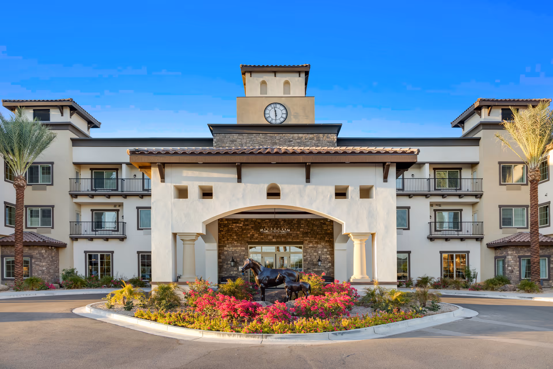 Front exterior view of The Park at Surprise Independent Living Community building with a clock tower, balconies, palm trees, and a landscaped roundabout featuring horse statues and colorful flowers under a clear blue sky.
