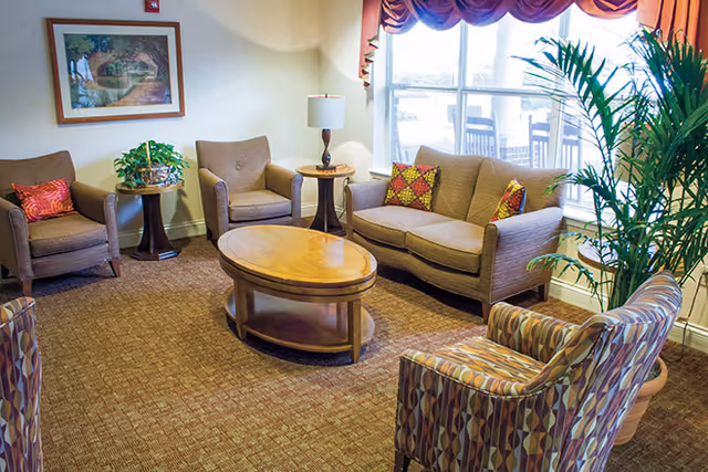 Well-lit seating area with a sofa, several armchairs, a round coffee table, side tables and plants by a large window.