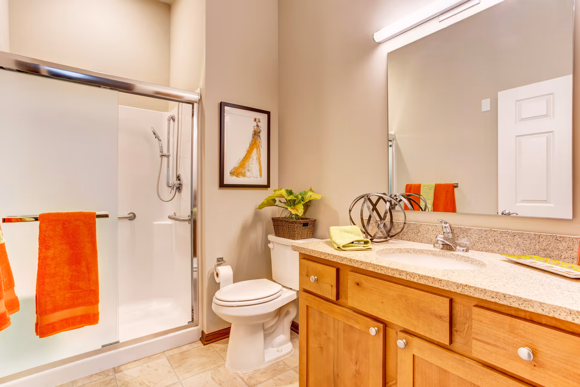A clean and well-lit bathroom featuring a glass-enclosed shower with a handheld showerhead, a white toilet with a plant on top, a wooden vanity with a granite countertop, a large mirror above the sink, and orange towels hanging on a towel rack.