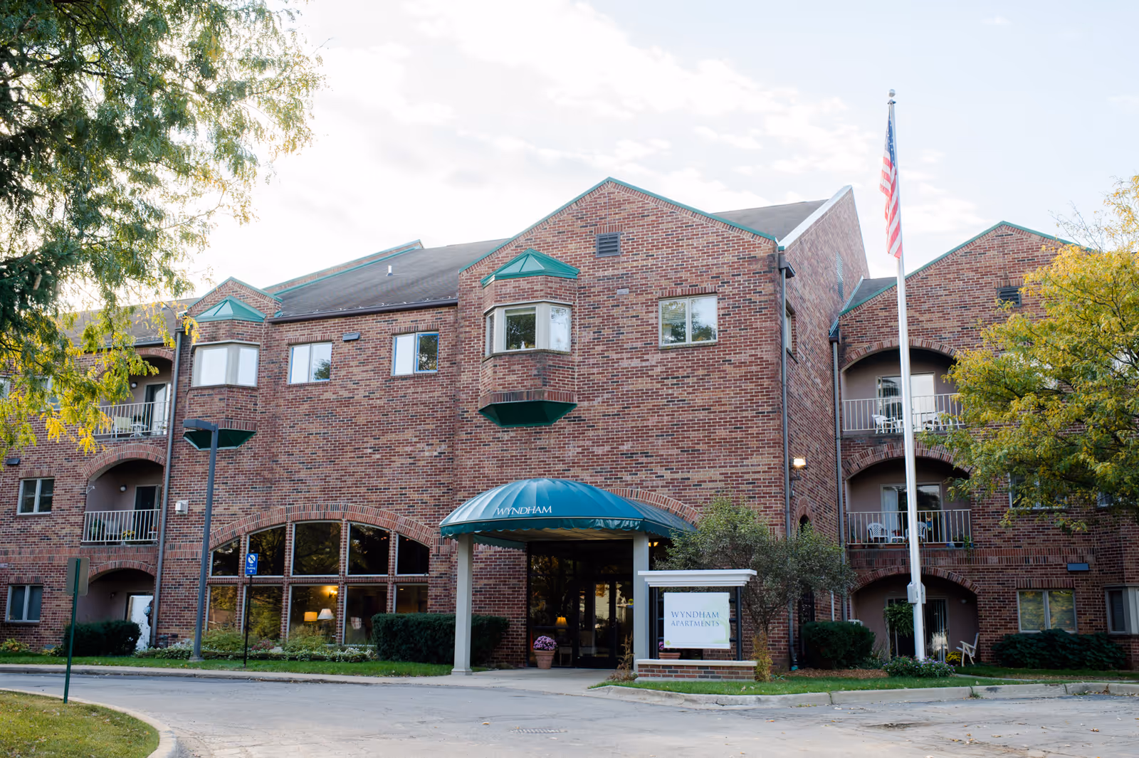 Exterior view of a three-story brick building with balconies, an entrance canopy labeled 'Wyndham', an American flag on a flagpole, and surrounding trees and landscaping.