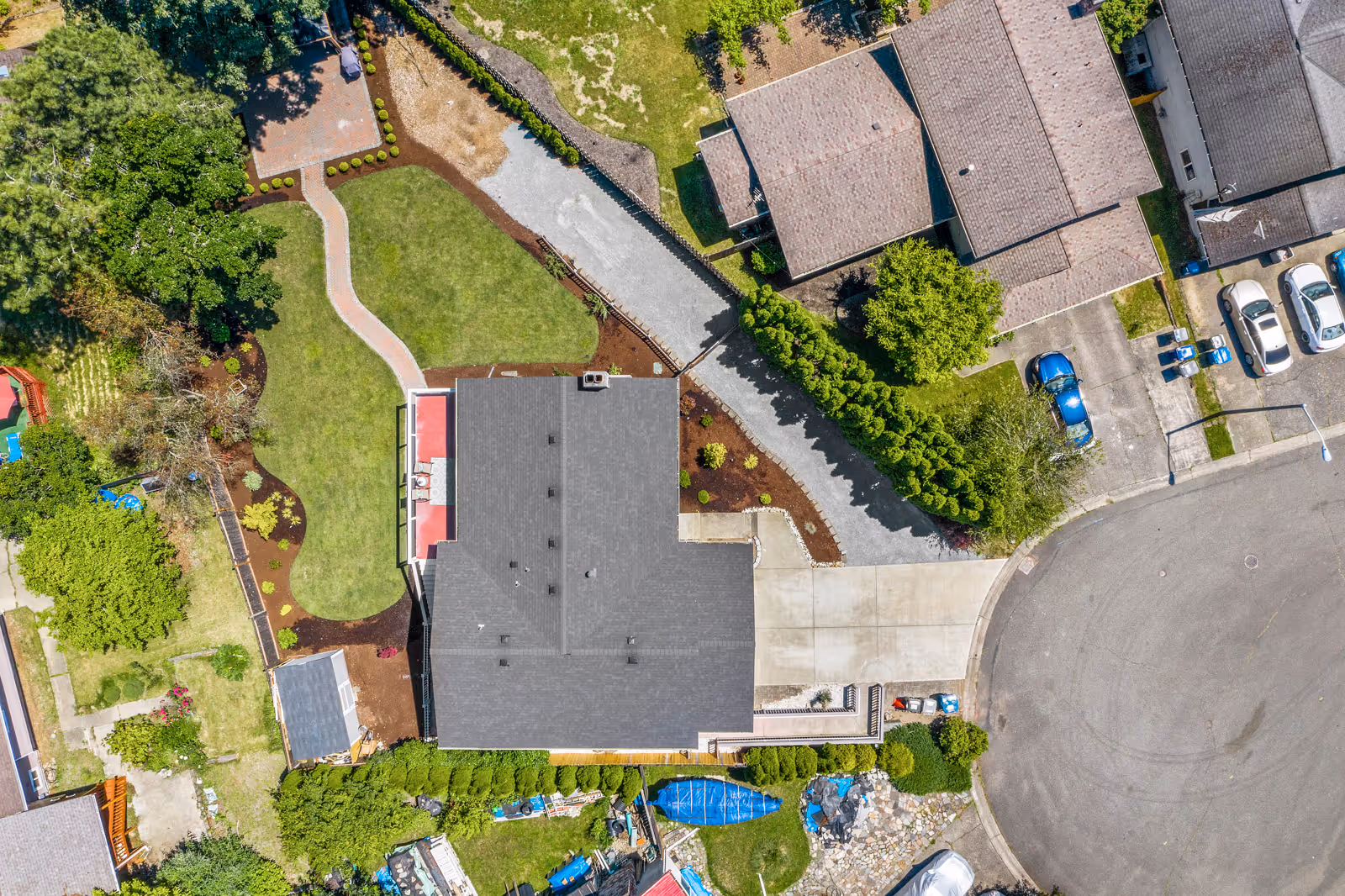 Aerial view of a residential property with a house, driveway, green lawn, garden areas, and surrounding neighboring houses. The property features a paved walkway, landscaped garden beds, and a cul-de-sac street with parked cars.