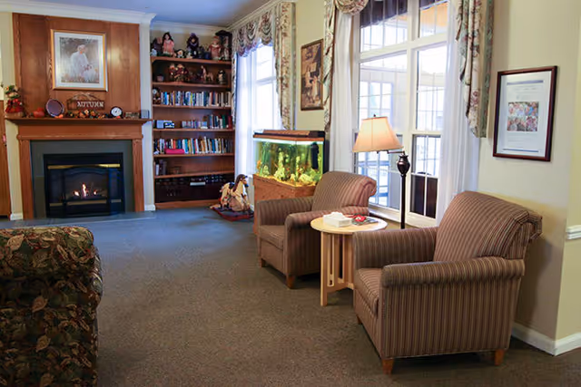 A cozy living room with two striped armchairs and a small round wooden table between them, a floor lamp, a large fish tank, a built-in wooden bookshelf filled with books and decorative items, and a fireplace with a wooden mantel. The room has large windows with floral curtains and framed pictures on the walls.