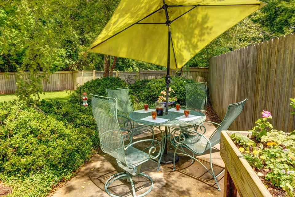 Outdoor patio area with a round metal table and four matching chairs under a large yellow umbrella. The table has several glasses filled with iced tea and napkins. Surrounding the patio are green bushes, a wooden fence, and a raised garden bed with flowers and plants.