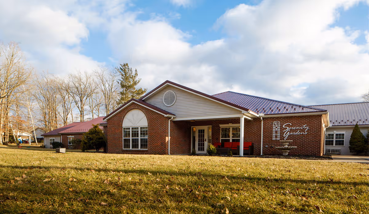 Front exterior of the single-story Serenity Gardens brick building with a grassy lawn and cloudy sky.
