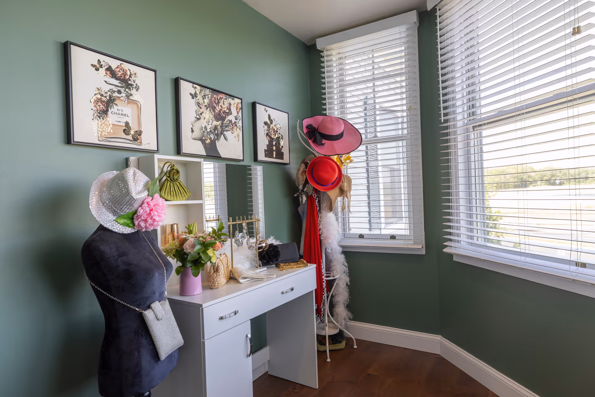 A bright corner room with green walls and large windows with white blinds. There is a white vanity table with a mirror, decorated with flowers and jewelry. A black mannequin dressed with a white hat and pink flower stands next to the vanity. On the wall above the vanity are three framed floral-themed artworks. A coat rack in the corner holds several hats and scarves.