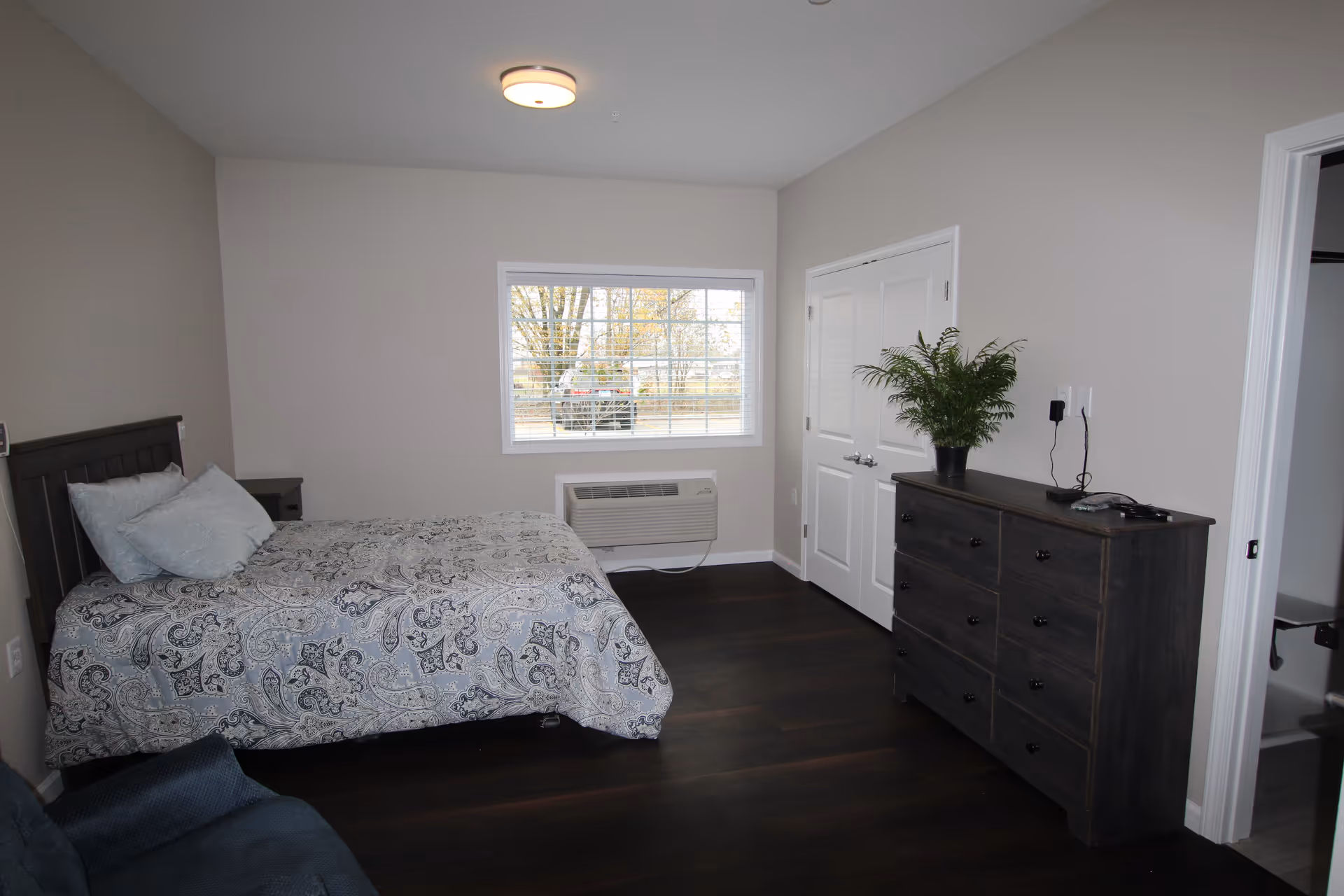 A bedroom with a bed covered in a patterned bedspread, two pillows, a dark wooden dresser with a potted plant on top, a window with blinds, and a door leading to a bathroom.