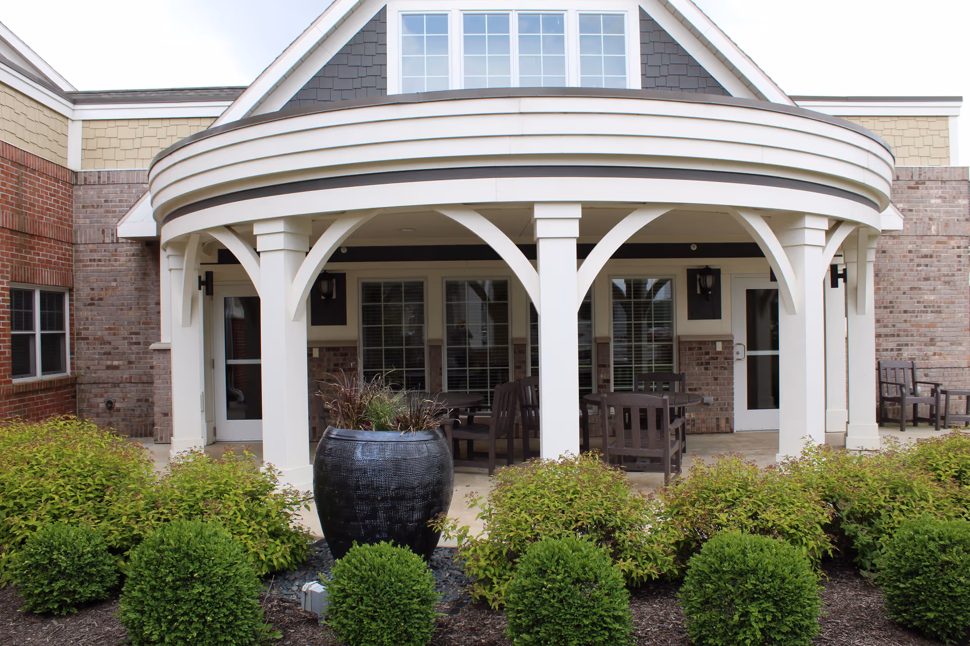 Front entrance porch with white arched columns, patio furniture, a large decorative planter, and landscaped shrubs.