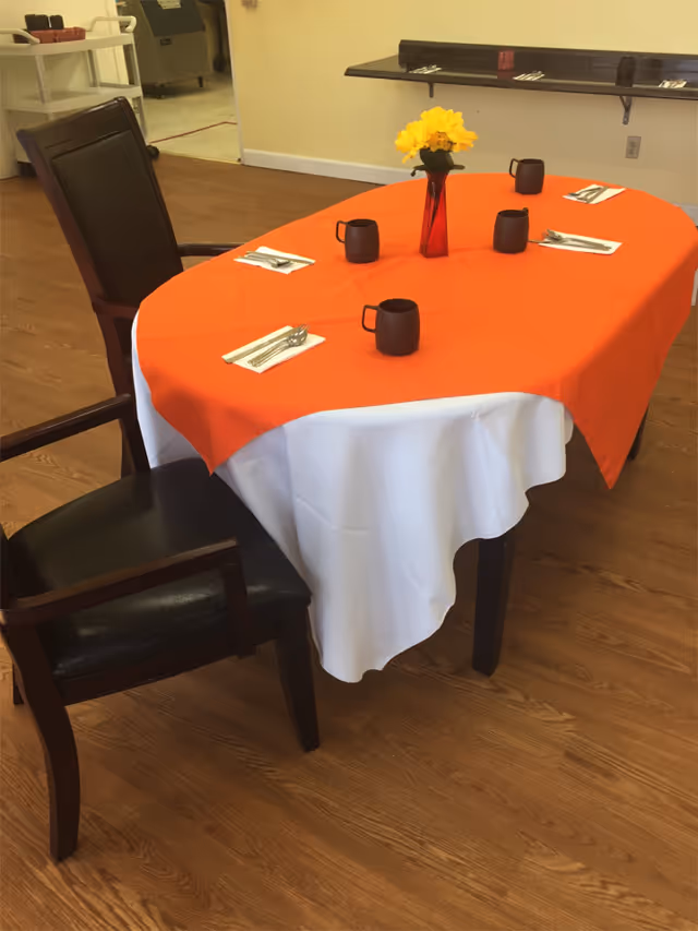 Dining table set with an orange tablecloth overlay, four mugs, utensils, and a vase of yellow flowers in a dining area.