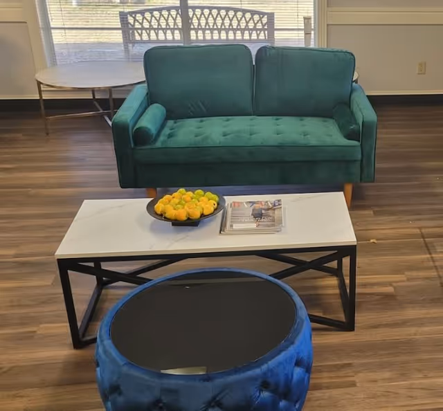 Small seating area with a green loveseat, a white coffee table holding a bowl of lemons and magazines, and a blue ottoman on wood flooring in front of windows.