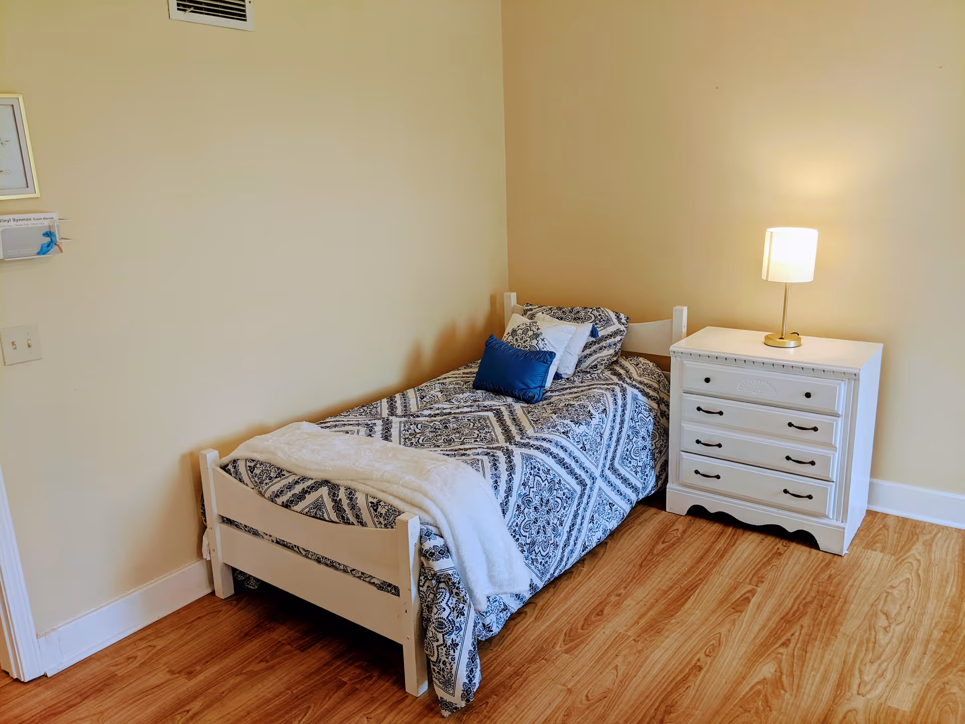 A small bedroom with a single white wooden bed featuring a blue and white patterned bedspread and pillows. Next to the bed is a white nightstand with four drawers and a lit table lamp on top. The room has light yellow walls and wood flooring.