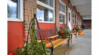 Covered exterior walkway of a brick senior living facility lined with decorative benches, planters and pumpkins.