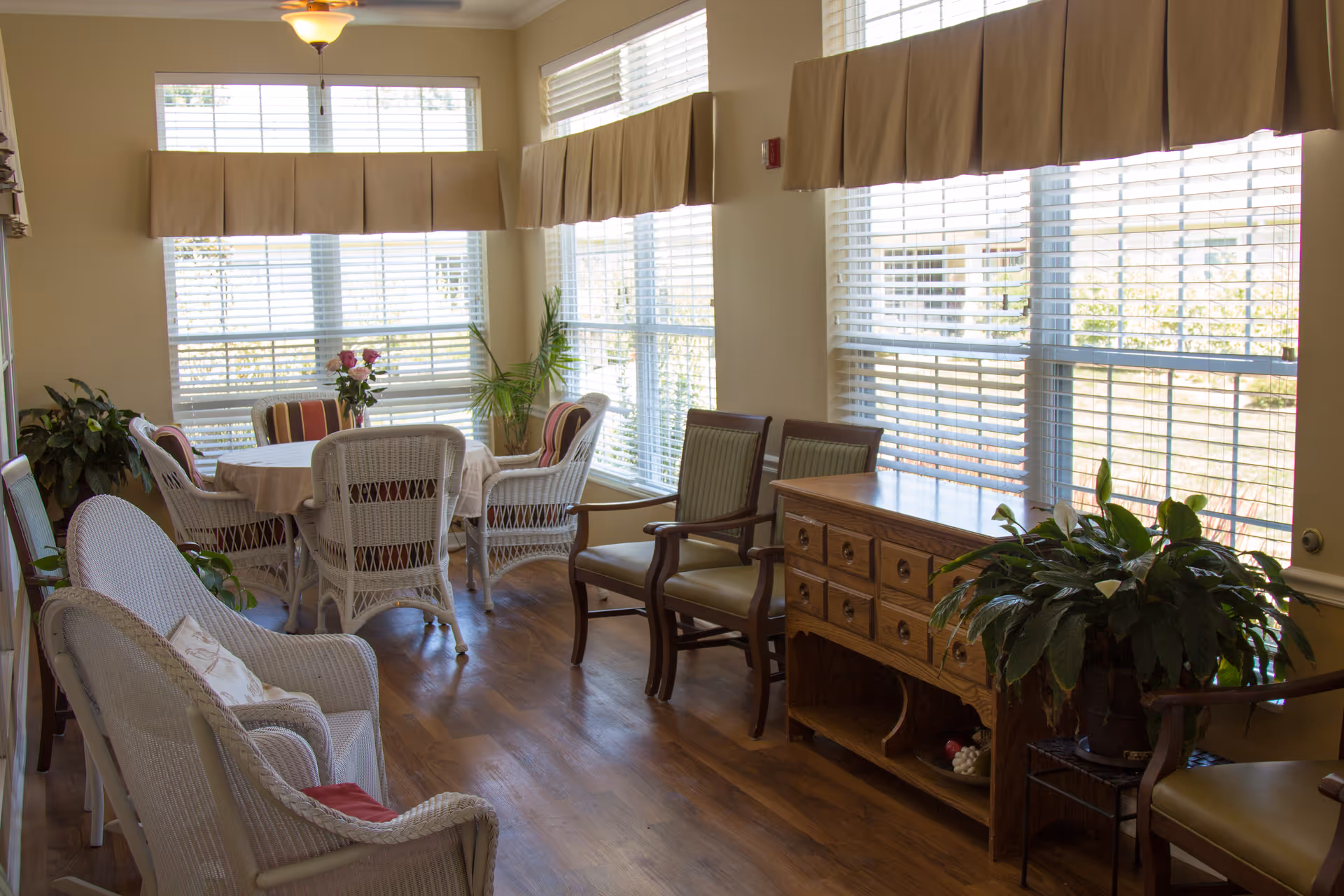Sunlit common room with wicker chairs, a round table and additional seating beside large windows.