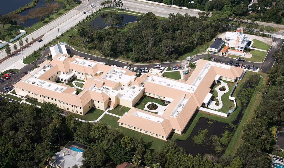 Aerial view of HarborChase of Sarasota, showing a large senior living facility with multiple connected buildings, surrounded by greenery, parking lots, and nearby roads.