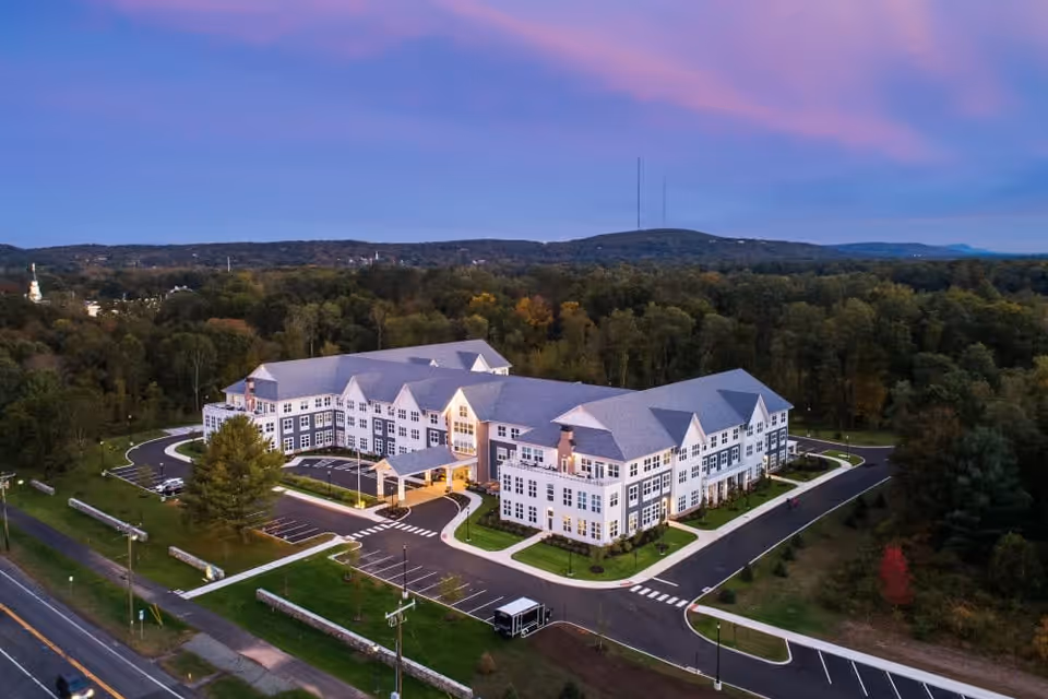 Aerial view of a large, modern senior living facility building named Riverbend at Farmington, surrounded by trees and greenery under a purple and blue evening sky. The building has multiple windows and a covered entrance with a driveway and parking spaces.