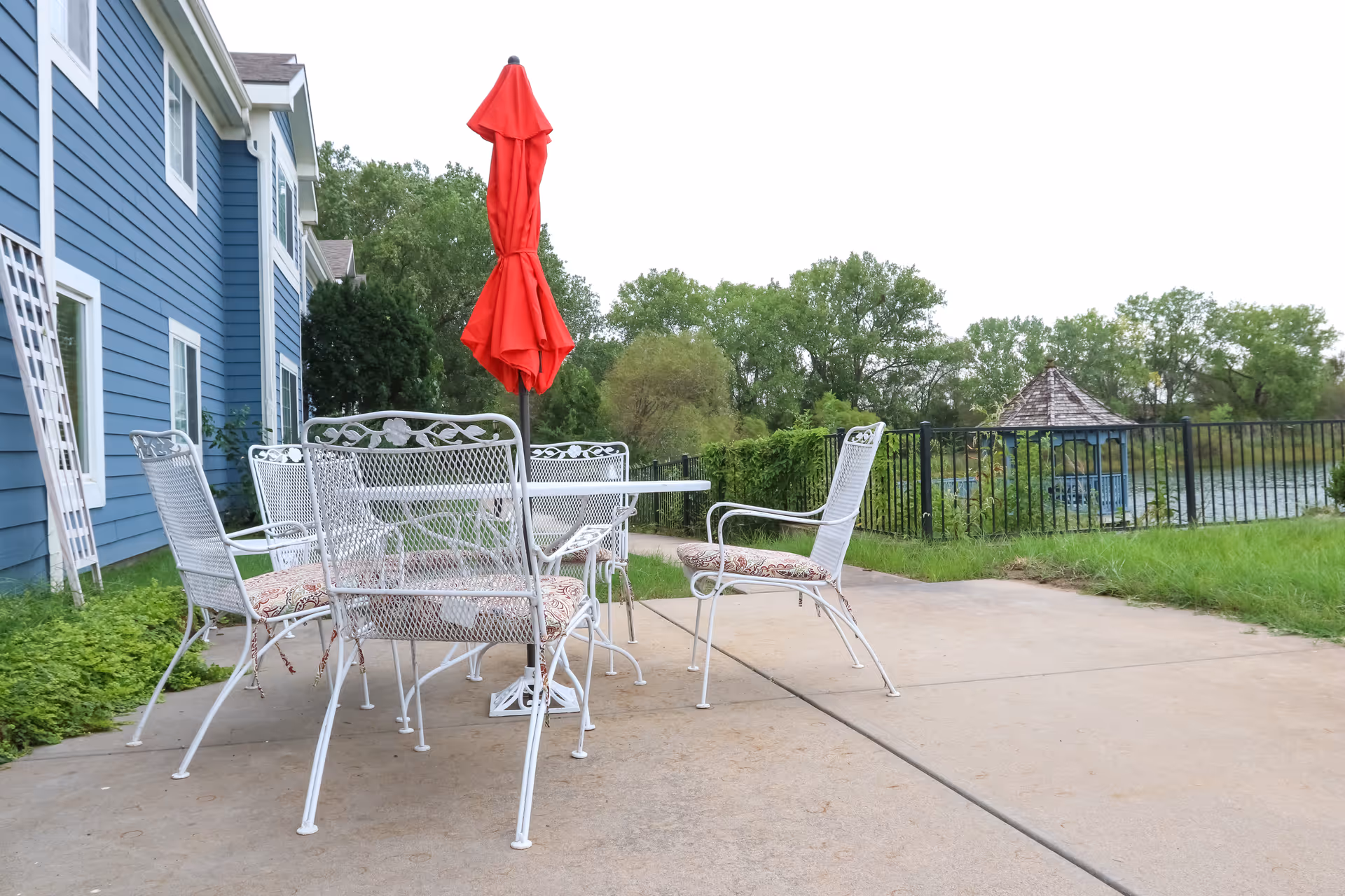 Outdoor patio area with white metal chairs and a table with a closed red umbrella, next to a blue building. In the background, there is a black metal fence, green trees, and a small gazebo near a body of water.