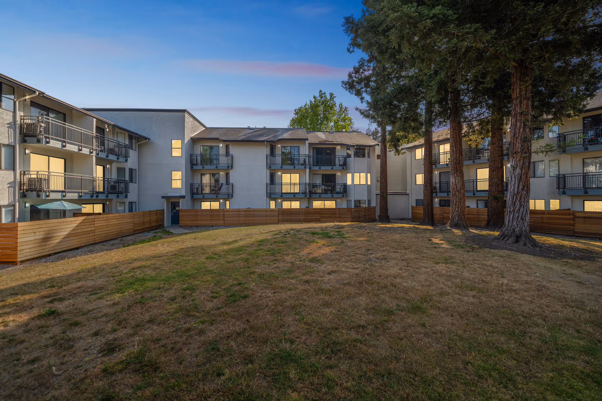 A grassy courtyard with tall trees surrounded by a three-story apartment building with balconies at dusk.