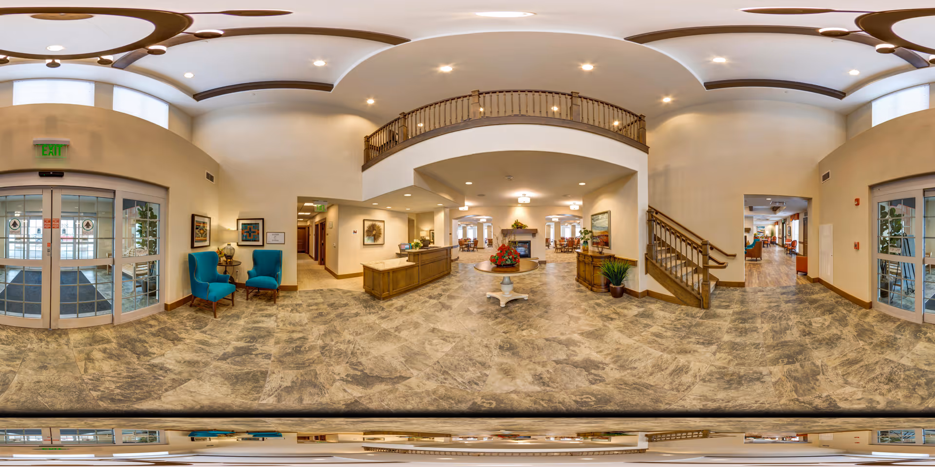 Wide panoramic view of a senior living facility lobby featuring a reception desk in the center, two blue armchairs with a small table on the left near glass entrance doors, a staircase on the right, and a round table with a flower arrangement in the middle. The space has high ceilings with recessed lighting and a balcony railing above the reception area. The floor is covered with stone-patterned tiles.