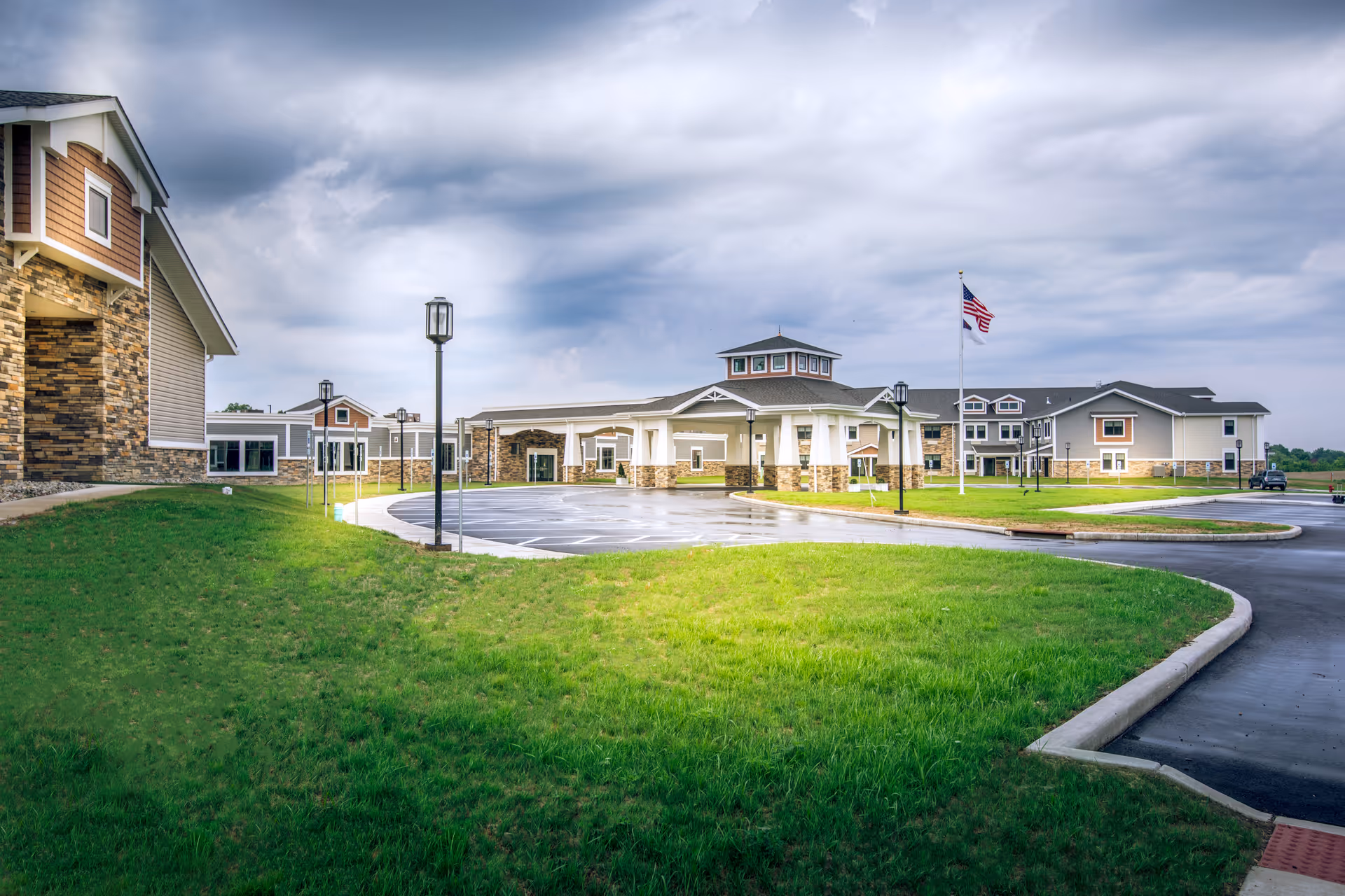 Exterior view of Shepherd of The Valley - Liberty senior living facility showing a large building with stone and beige siding, a circular driveway, green lawn, several lamp posts, and an American flag on a flagpole under a cloudy sky.