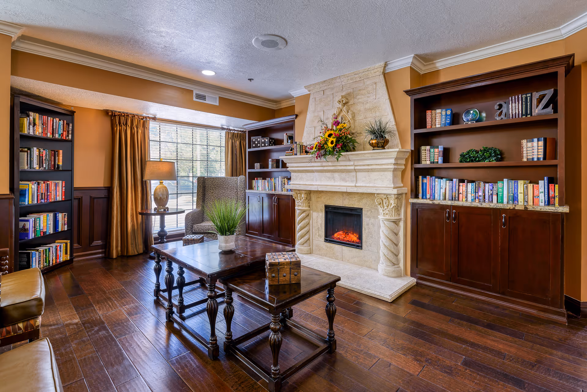 A cozy living room with a stone fireplace in the center, decorated with a floral arrangement and plants on the mantel. The room features dark wood bookshelves filled with books on either side of the fireplace, a large window with golden curtains, a patterned armchair, a wooden coffee table with a small plant and decorative box, and hardwood flooring.