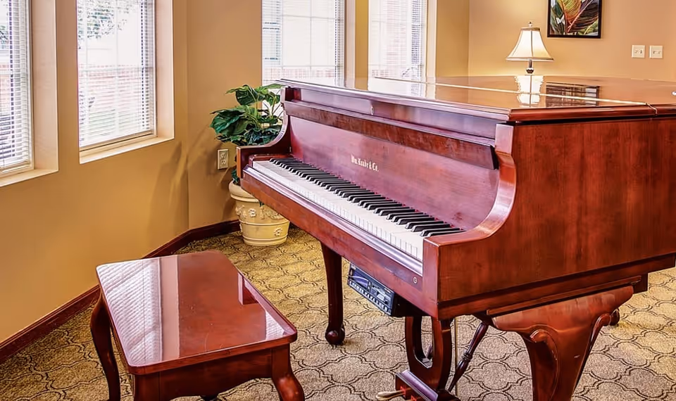 Mahogany grand piano and matching bench in a sunlit common room with windows and a potted plant.