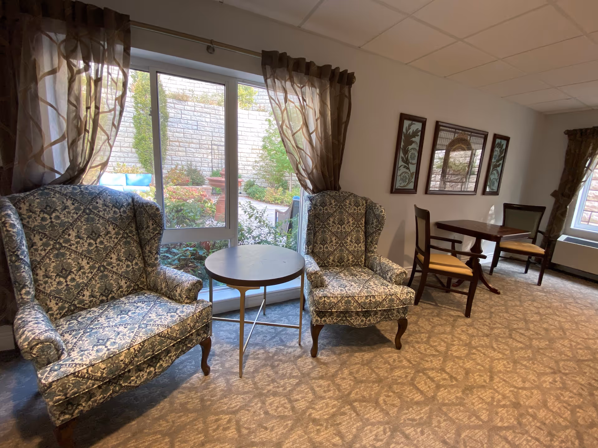 A cozy sitting area in a senior living facility with two patterned armchairs separated by a small round table. Behind the chairs is a large window with sheer curtains, showing a view of a garden with a stone wall and greenery. To the right, there is a wooden table with two chairs and framed artwork on the wall.