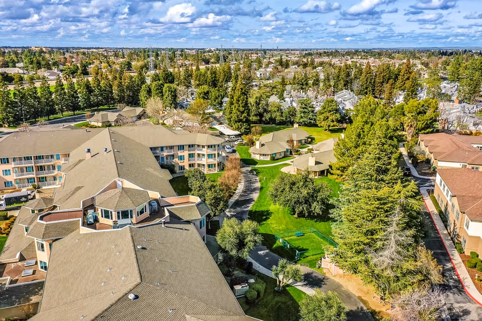 Aerial view of Solstice Senior Living at Bakersfield showing multiple residential buildings with beige roofs surrounded by green lawns, trees, and pathways under a partly cloudy sky.