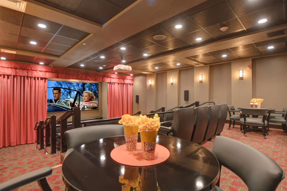 Interior view of a cozy movie theater room with rows of comfortable black seats facing a large screen showing a classic movie scene. The room has red carpet with a floral pattern, beige walls with wall sconces, and a ceiling with recessed lighting. In the foreground, there is a round table with two popcorn containers and chairs around it.