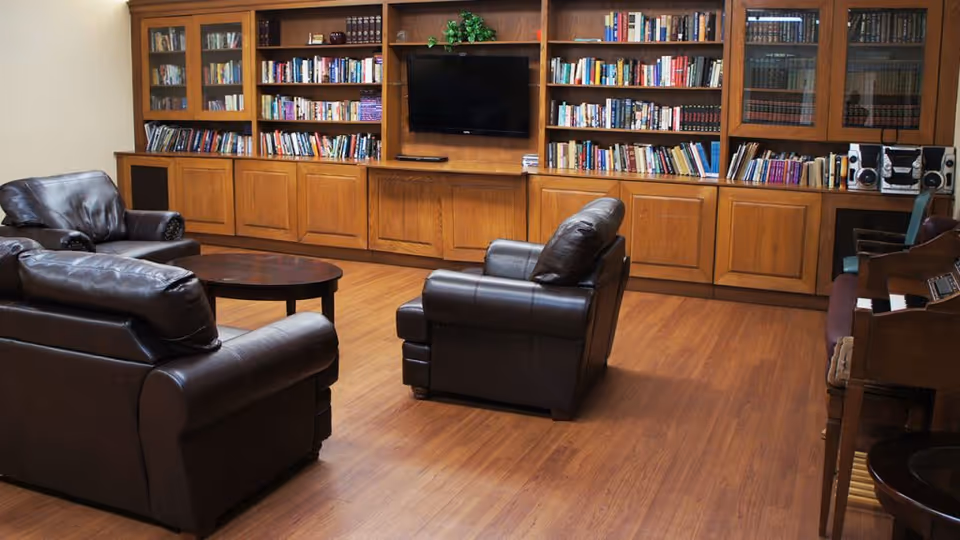 A cozy common area with dark leather armchairs and a sofa arranged around a wooden coffee table. Behind them is a large wooden bookshelf filled with books and a flat-screen TV mounted in the center. The room has wooden flooring and a piano with a stereo system on the right side.