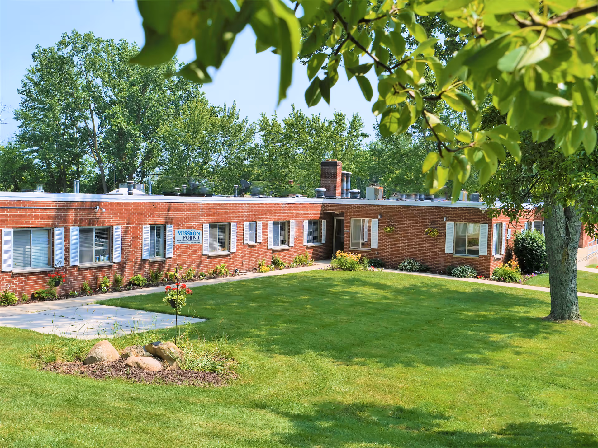 Single-story brick building with multiple windows and a sign that reads 'MISSION POINT' surrounded by a well-maintained green lawn, trees, and some landscaping with rocks and flowers under a clear blue sky.