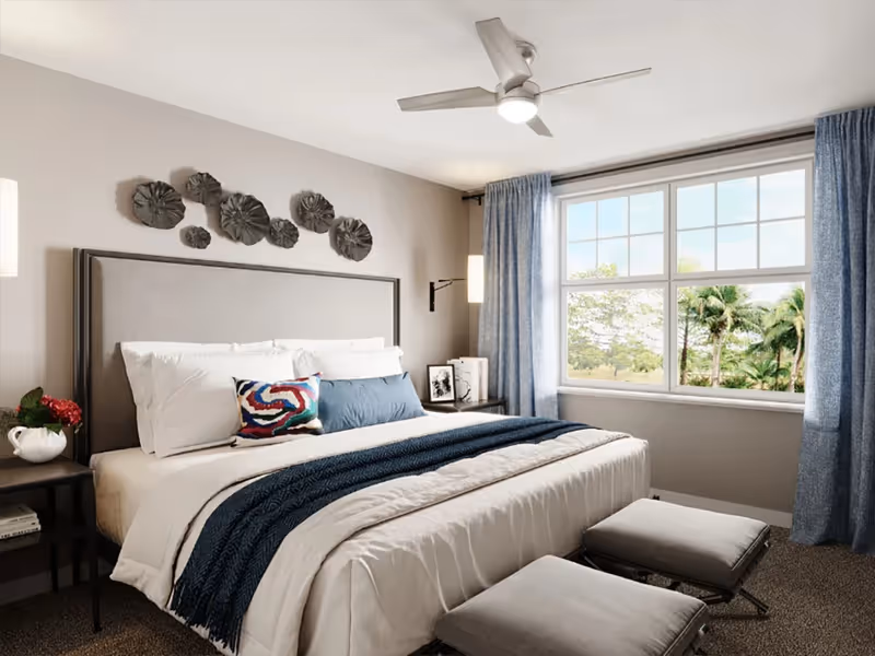 A bright and cozy bedroom with a large bed featuring white and blue bedding, decorative pillows, and a gray upholstered headboard. Two gray ottomans are placed at the foot of the bed. The room has a large window with blue curtains, letting in natural light and showing a view of trees outside. Wall-mounted lamps and a ceiling fan with a light fixture are also visible.