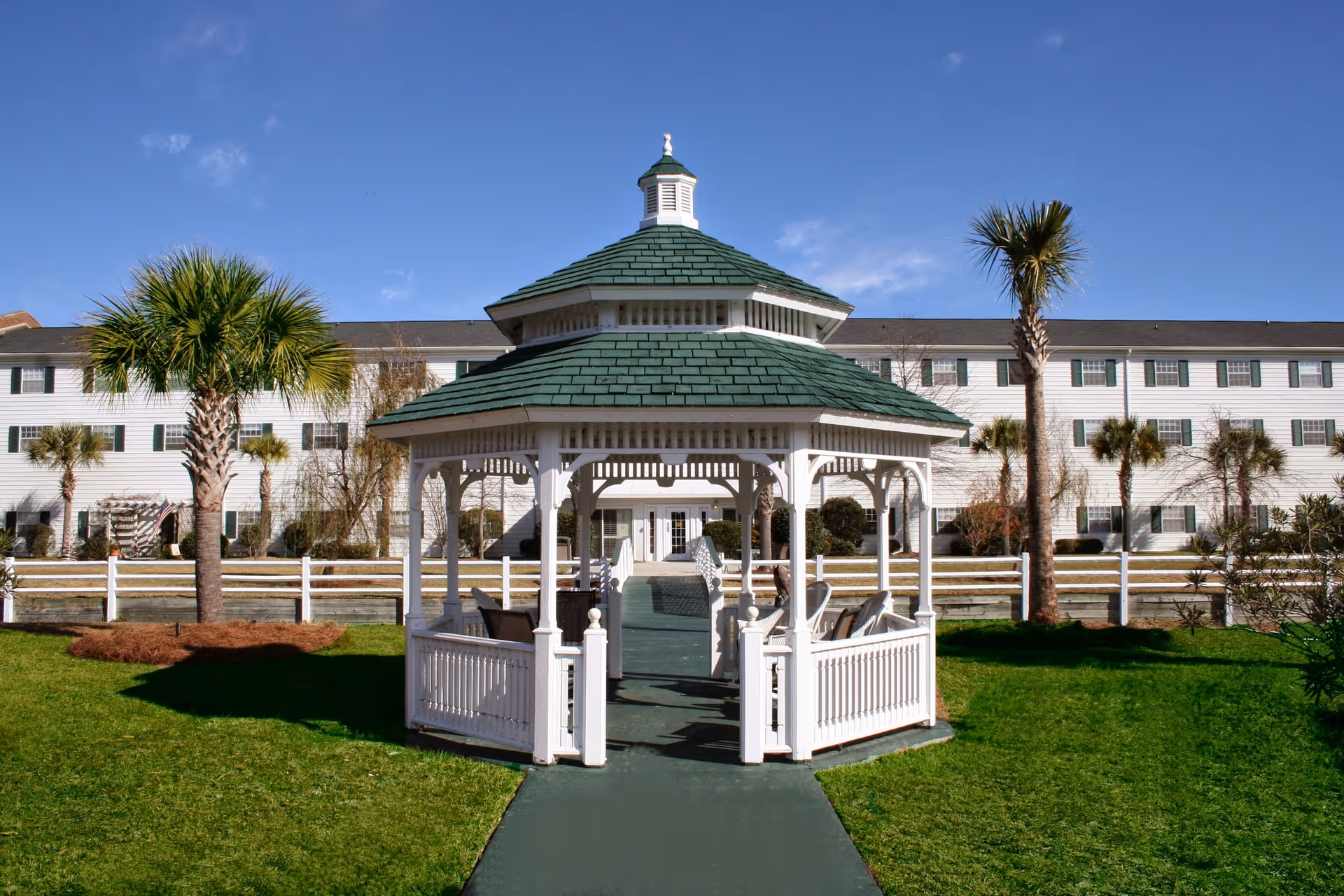 A white gazebo with a green roof situated on a green lawn with palm trees around it. In the background, there is a large white multi-story building with green shutters and a white fence in front of it under a clear blue sky.