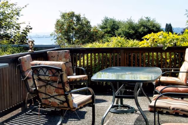 Outdoor balcony patio with cushioned chairs, glass table, and greenery.