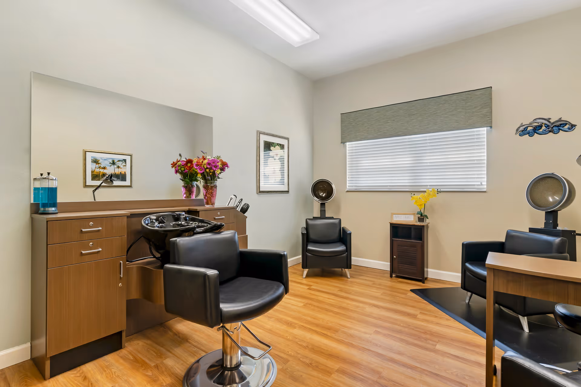 Interior of a salon room with wooden flooring, a large mirror above a wooden cabinet with a black salon sink and chair in front. There are two black salon chairs with hair dryers behind them, a small cabinet with yellow flowers, and framed artwork on the walls. A window with blinds and a green valance is on the right wall.