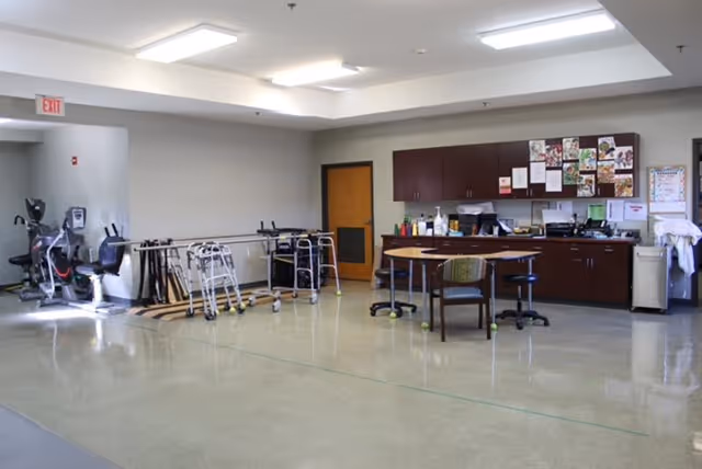 A spacious room in a nursing care center with exercise equipment including stationary bikes and walkers lined up against the wall. There is a wooden table with chairs in the center and cabinets with various supplies and bulletin boards on the wall behind it. The floor is shiny and the ceiling has recessed lighting.