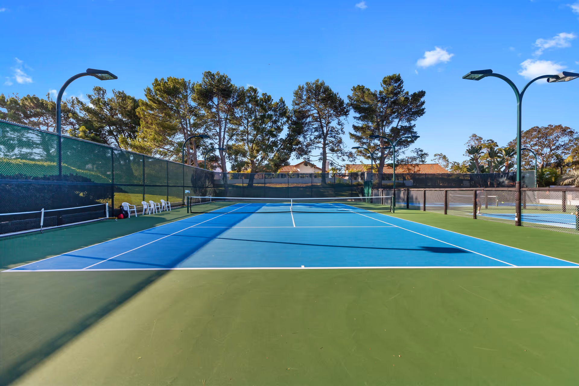 Outdoor tennis court with blue and green surface, nets, surrounding fences and lights, with trees and a clear sky in the background.