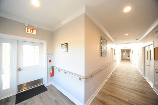A bright, clean hallway in a senior living facility with light wood flooring, beige walls, handrails along the walls, and an exit door with a lit exit sign above it. There is a fire extinguisher mounted on the wall near the door and framed artwork hanging on the walls.