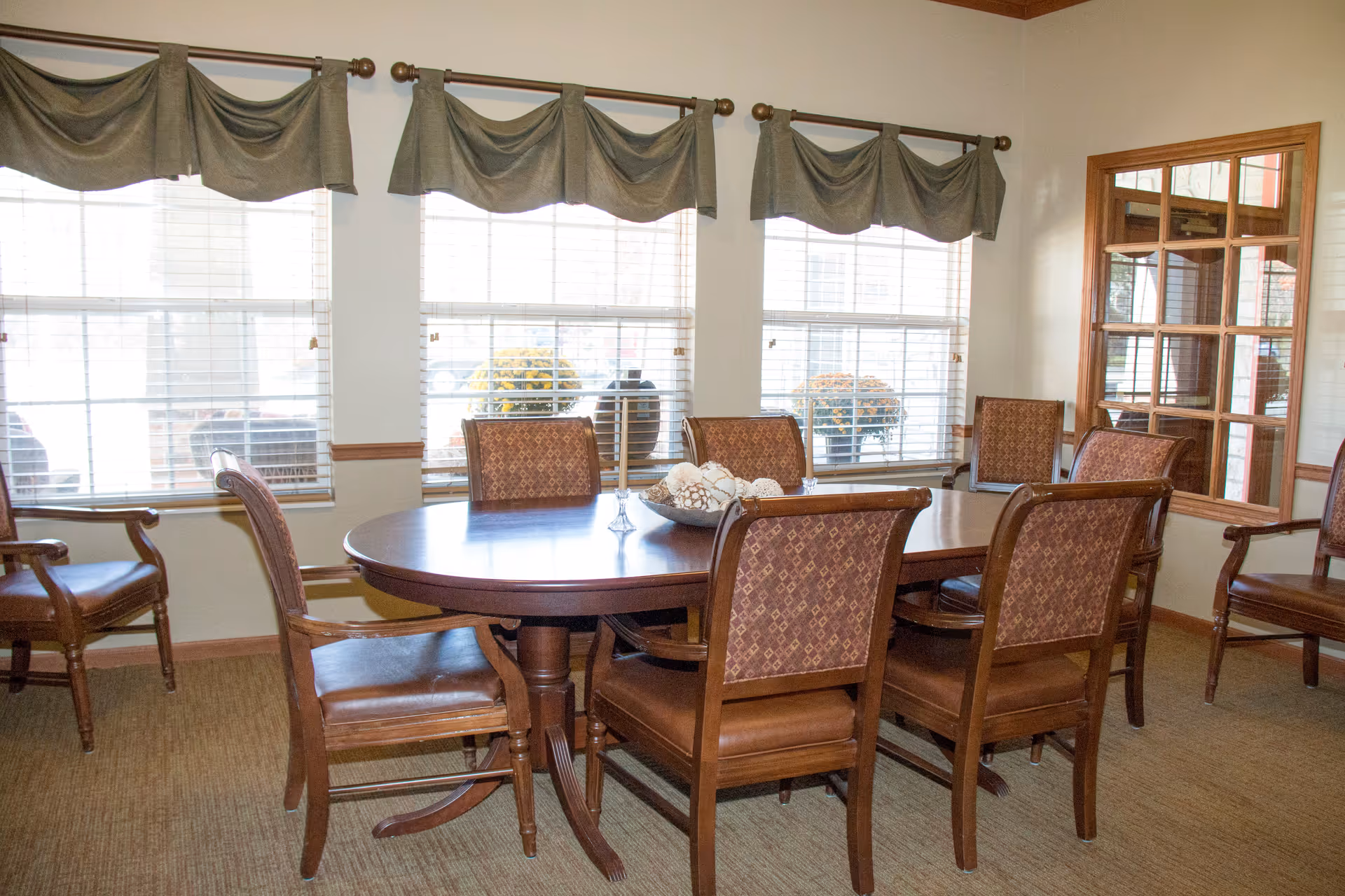 A dining room with a large wooden oval table surrounded by upholstered chairs in front of three windows with green valances.