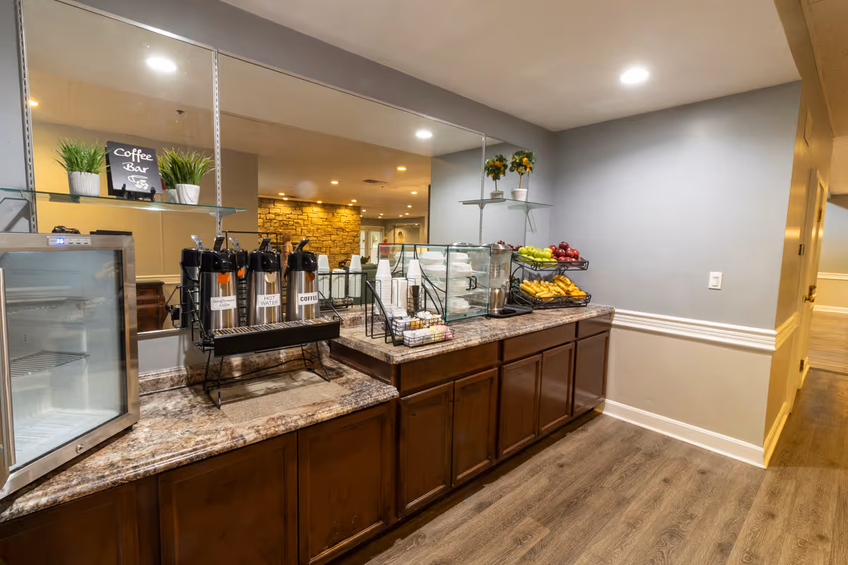 A coffee bar area in a senior living facility with a countertop holding coffee dispensers labeled Decaf, Hot Water, and Coffee, disposable cups, plates, and baskets of fruit including bananas, apples, and grapes. The background features a mirror reflecting a dining area with tables and chairs, and the floor is wood with beige walls.