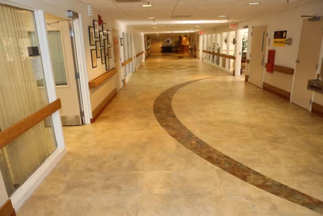 A wide, well-lit hallway in a healthcare or rehabilitation facility with beige tiled floors featuring a curved darker tile pattern. The hallway has handrails on both sides, several doors, framed pictures on the walls, and an exit sign visible at the far end.