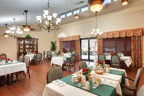 A bright and spacious dining room in an assisted living facility with multiple tables covered in white and green tablecloths, set with glasses, cups, and napkins. The room features wooden flooring, decorative chandeliers, large windows with floral curtains, and a wooden cabinet displaying dishes and glassware.