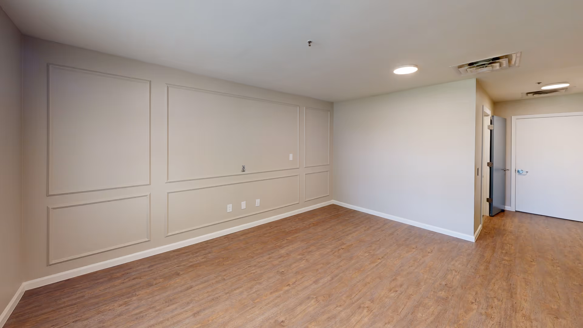 Empty room with light beige walls featuring decorative molding, wood-look flooring, a white door at the far end, and ceiling lights.