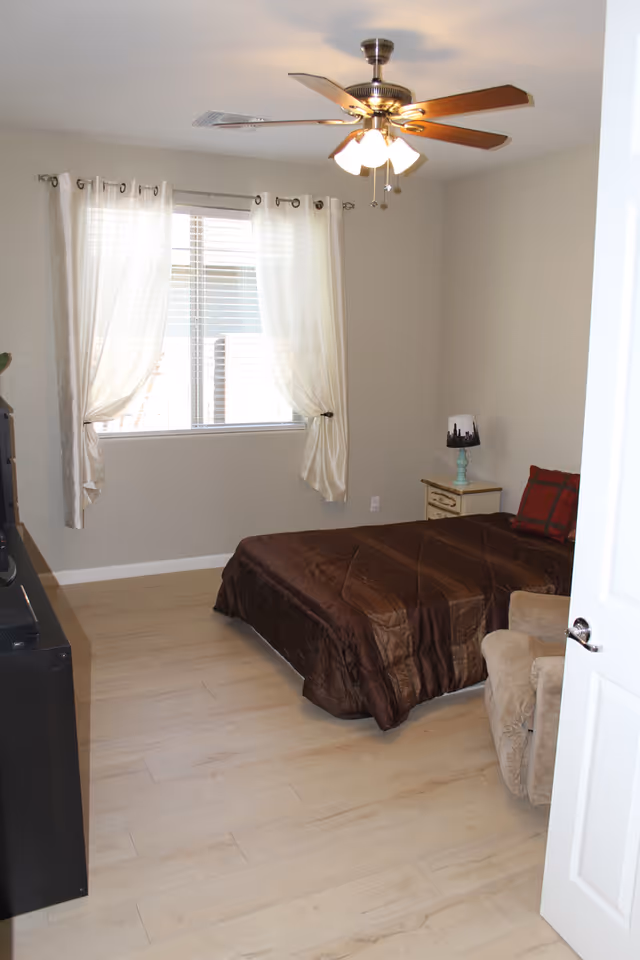 A small bedroom with a single bed covered in a brown comforter and a red pillow. There is a beige armchair next to the bed, a small nightstand with a lamp featuring a cityscape design, a window with white curtains, and a ceiling fan with lights. The floor is light-colored wood, and the walls are painted light gray.